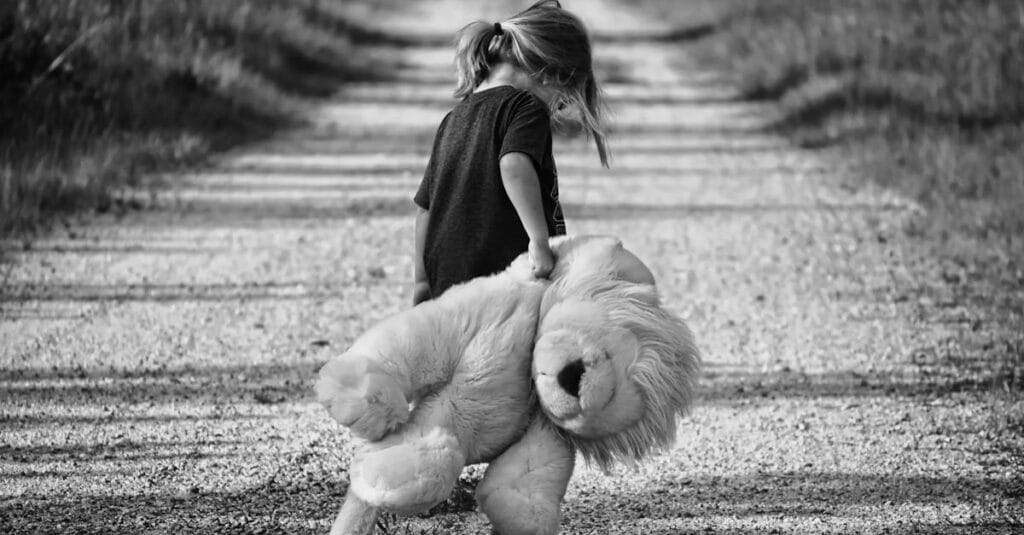 Monochrome image of a child holding a teddy bear while walking down a dirt road, conveying a sense of solitude.