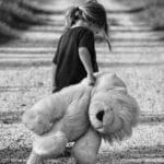 Monochrome image of a child holding a teddy bear while walking down a dirt road, conveying a sense of solitude.