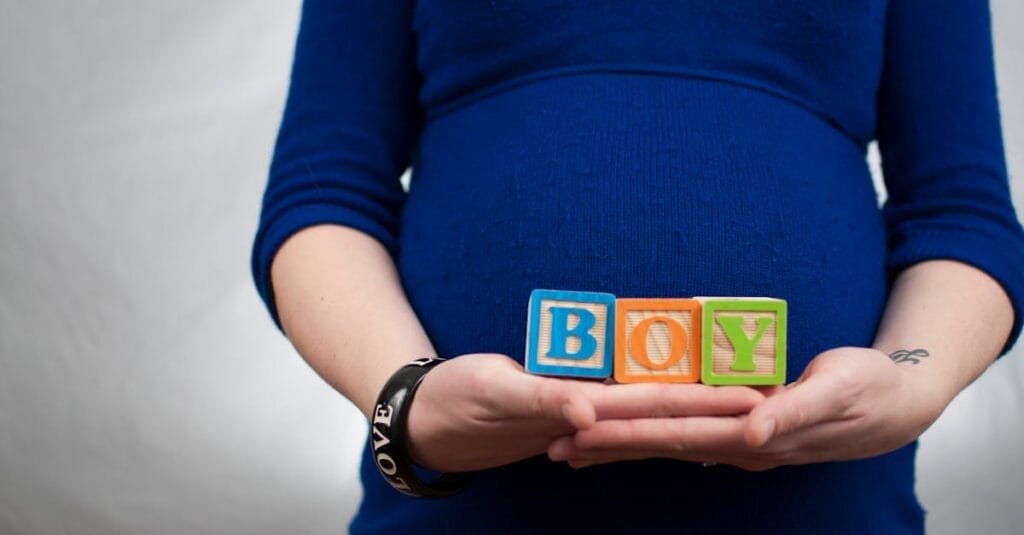 Pregnant woman in blue dress holds 'boy' blocks, symbolizing a baby boy announcement.