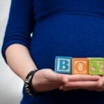 Pregnant woman in blue dress holds 'boy' blocks, symbolizing a baby boy announcement.