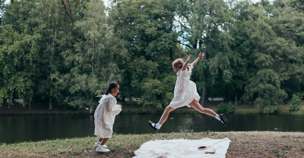 Two girls in white dresses playing joyfully by a lake in a lush green park.