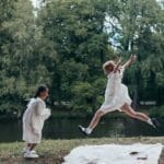 Two girls in white dresses playing joyfully by a lake in a lush green park.