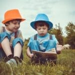 Two young boys enjoying a book together outdoors on a sunny day, wearing vibrant hats.