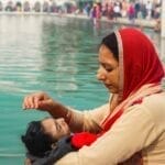 A mother comforts her child by the serene waters, wearing a red headscarf.