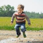 A cheerful child in a striped sweater jumps into a puddle on a sunny day in Czechia.