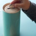 A child placing a coin into a teal piggy bank, representing saving money and financial education.