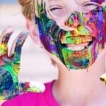 A joyful child with vibrant face paint and hands, expressing happiness and creativity.