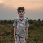 A contemplative boy stands alone in a grassy field at sunset, exuding calm.