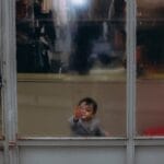 A young child peers through a glass window, touching the pane with curiosity.