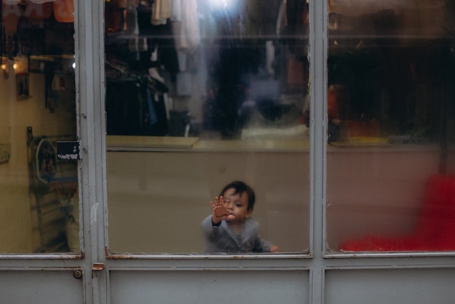 A young child peers through a glass window, touching the pane with curiosity.