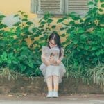 A young girl sits on a bench outdoors, immersed in a book with lush greenery around her.