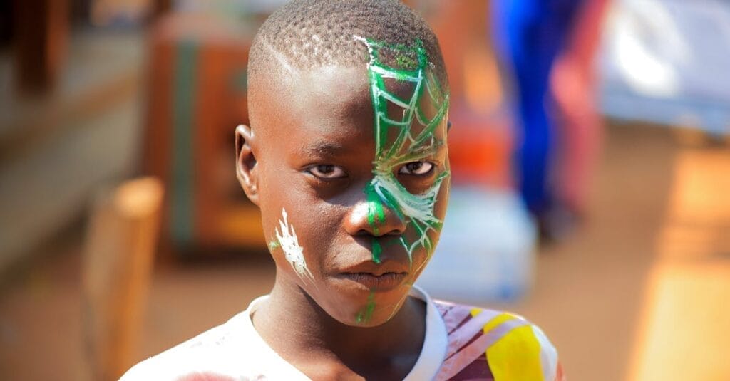 A young boy with intricate Spider-themed face paint poses outdoors in bright lighting.