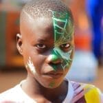 A young boy with intricate Spider-themed face paint poses outdoors in bright lighting.