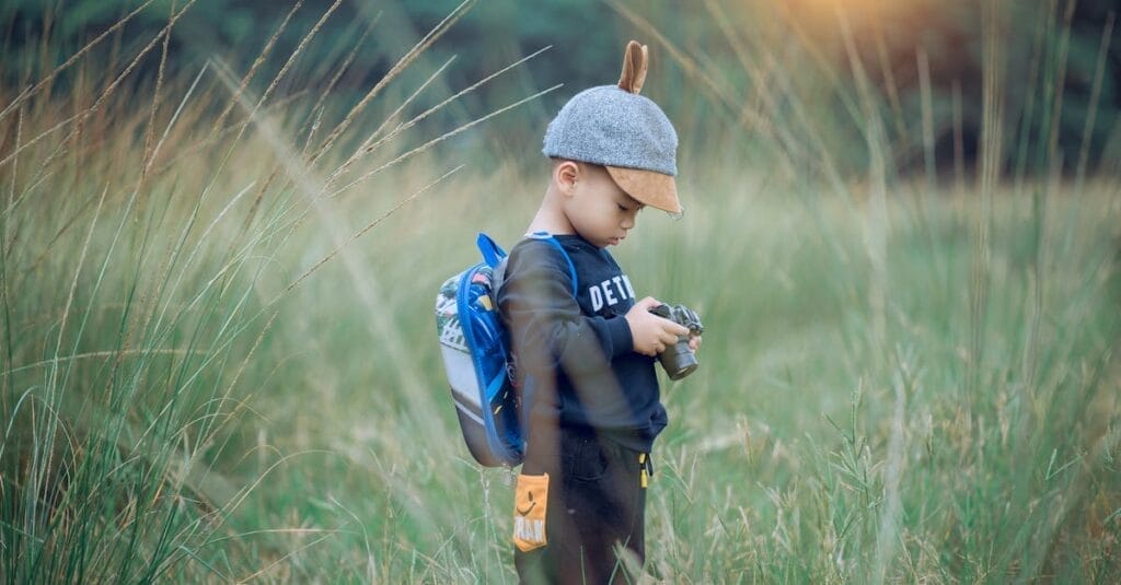 A young boy wearing a cap explores a grassy field with a camera, enjoying outdoor adventure.