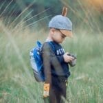 A young boy wearing a cap explores a grassy field with a camera, enjoying outdoor adventure.