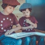 Two young boys wearing hats and star shirts reading a book outdoors on a wooden porch.