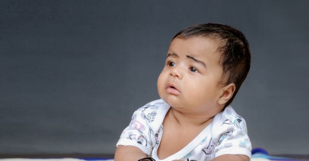Cute baby boy in a white outfit lying on a colorful blanket with a gray background.