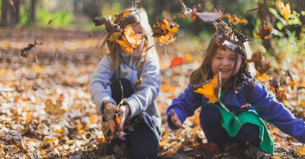 Two smiling girls playing joyfully amidst colorful fall leaves in a sunlit forest