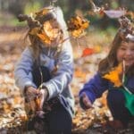 Two smiling girls playing joyfully amidst colorful fall leaves in a sunlit forest