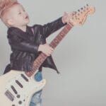 Stylish young boy with a mohawk strumming an electric guitar, wearing a leather jacket and blue jeans.