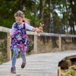 Young girl in a floral dress runs with toy rabbit on a forest boardwalk, embracing nature.