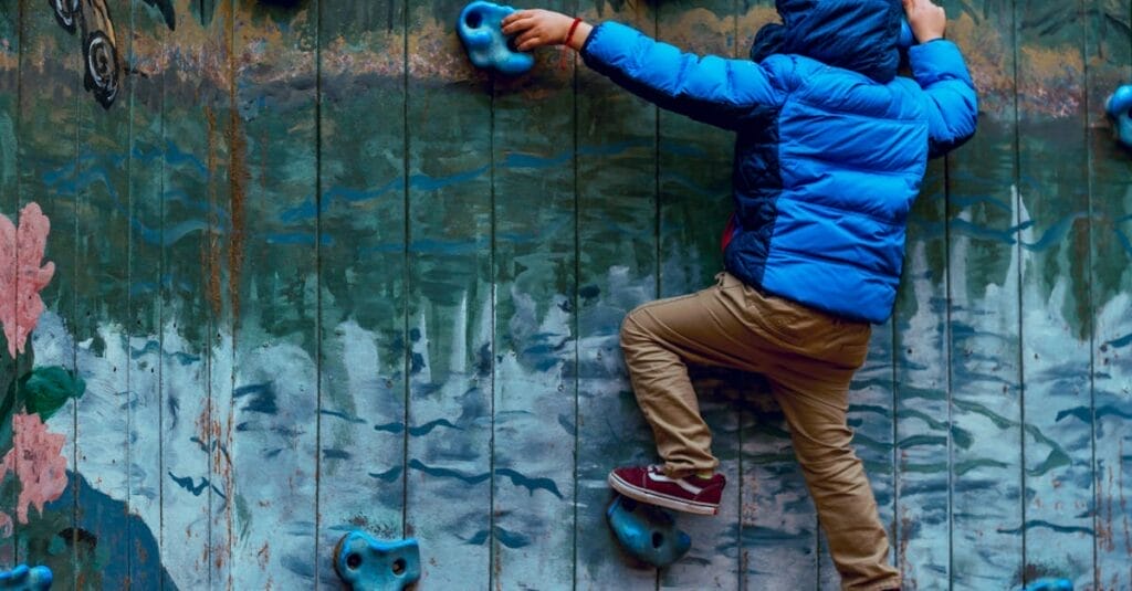 Young boy in blue jacket climbing a painted rock wall outdoors, showcasing adventure and play.