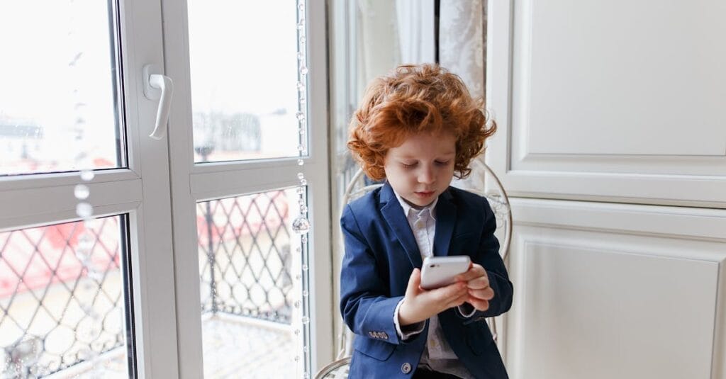 Red-haired boy wearing a suit using a smartphone indoors, seated near a window.