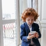 Red-haired boy wearing a suit using a smartphone indoors, seated near a window.