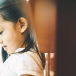 A serene portrait of a young girl in profile, thoughtfully looking down in a warmly lit indoor setting.