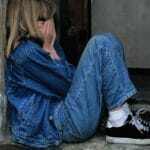 A young girl in denim sits alone in a doorway looking distressed, conveying loneliness.