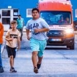 Children running in an urban setting with police motorcycle escort and emergency vehicles present.