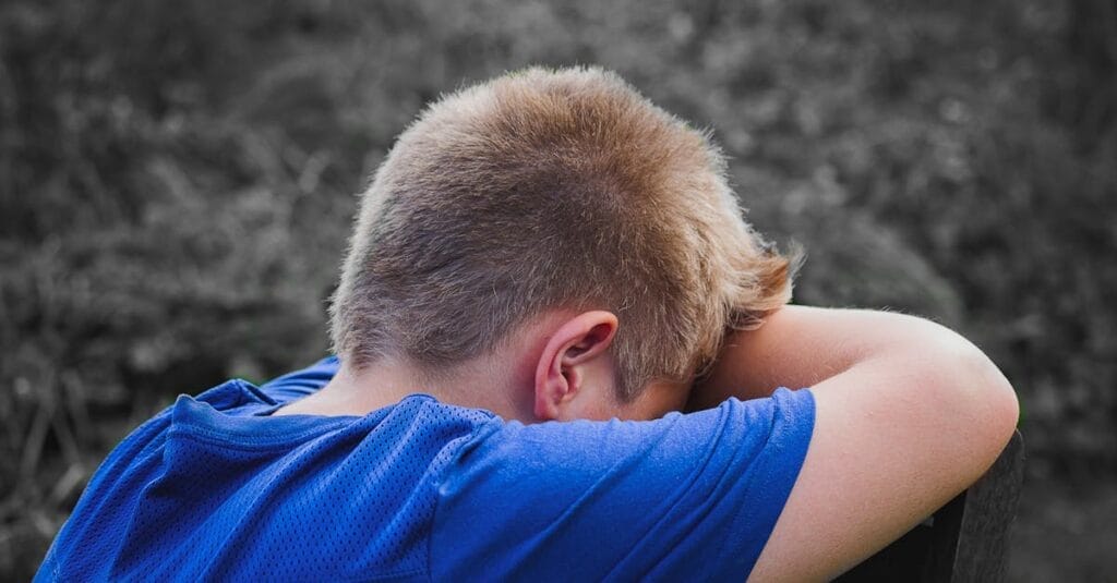 A young boy in a blue shirt rests his head on his arms outdoors, appearing sad and contemplative.