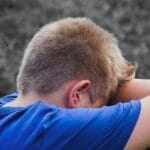 A young boy in a blue shirt rests his head on his arms outdoors, appearing sad and contemplative.