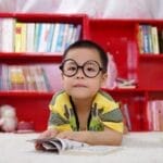 A cute child in glasses reading a book, surrounded by colorful shelves. Perfect shot of childhood learning indoors.