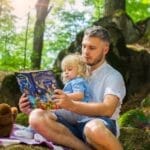 Father and toddler bonding over a book in a sunny forest setting.