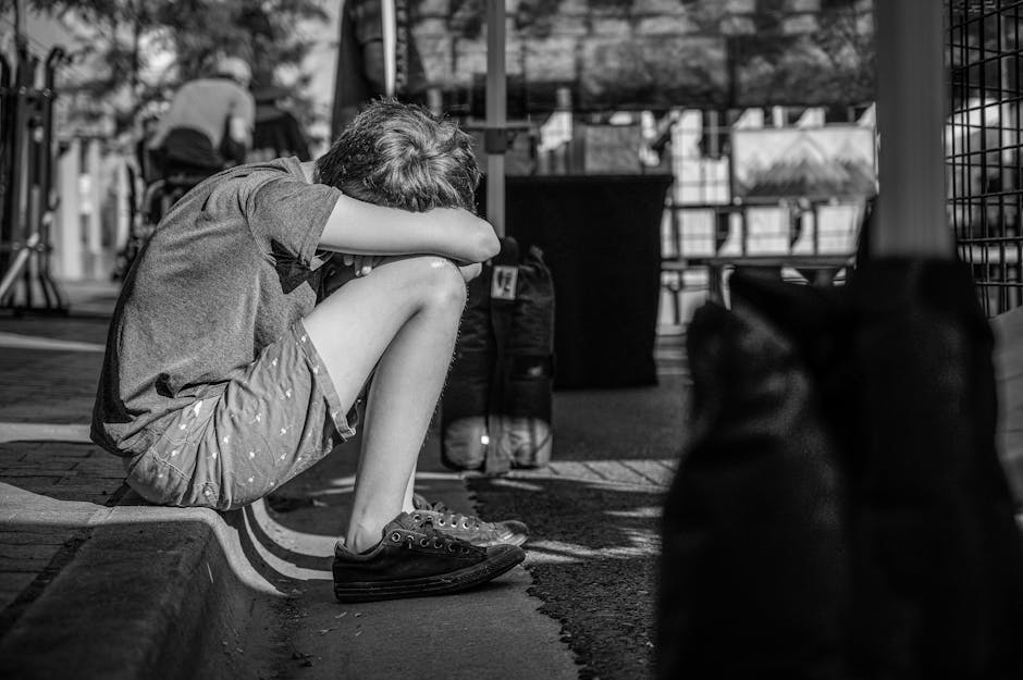 A child sitting alone on a curb, depicting solitude and contemplation in a monochrome city setting.