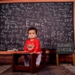 Cute baby in a classroom setting with a large chalkboard filled with equations.