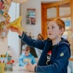 A young boy enjoying a creative activity with a paper airplane in a lively classroom setting.