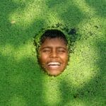 A child's joyful face emerges from a green duckweed-covered pond, creating a delightful scene in nature.