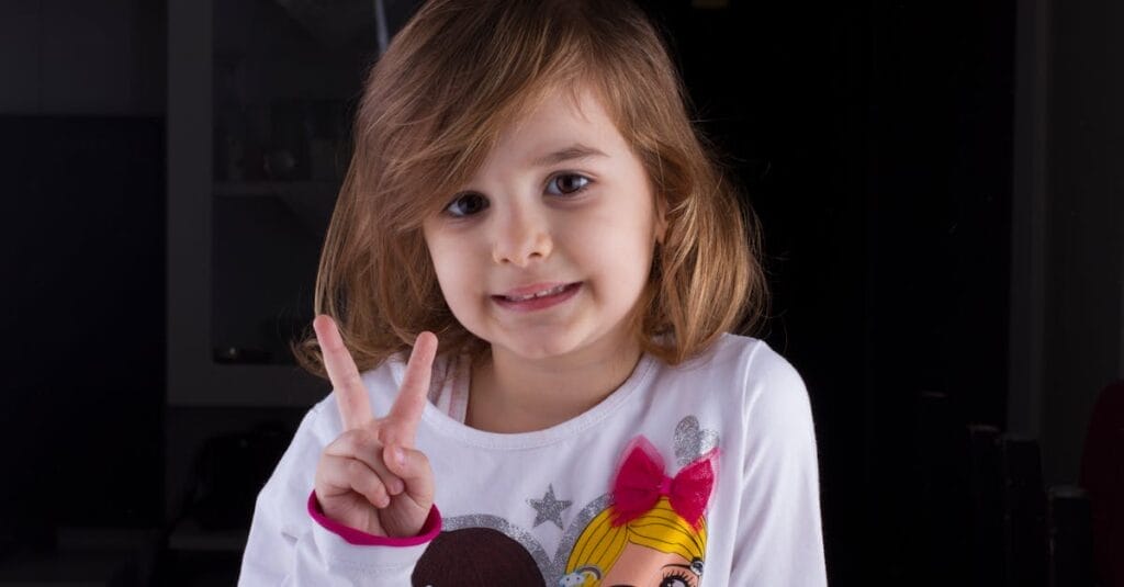 Child smiling indoors while making a peace sign with fingers, wearing playful attire.
