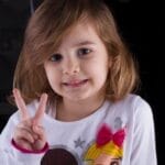 Child smiling indoors while making a peace sign with fingers, wearing playful attire.