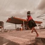 A young girl runs past a gasoline station under a cloudy sky in an urban environment.