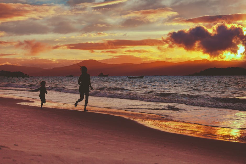 Silhouettes of a child and adult running along a beach at sunset with a dramatic sky and gentle waves.
