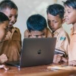 A group of young boys in school uniforms collaborating over a laptop in a classroom setting.