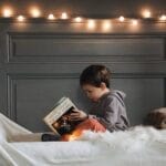 A young boy reads a book in a cozy bedroom setting with warm string lights and soft textures.