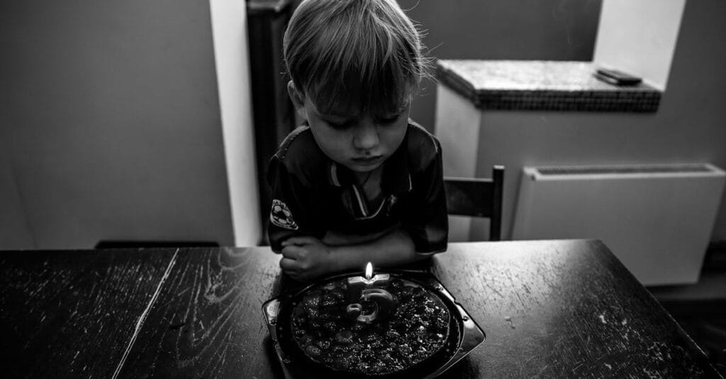 A young boy contemplatively looks at a birthday cake with a single candle, captured in black and white.