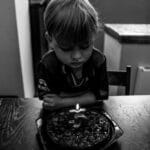 A young boy contemplatively looks at a birthday cake with a single candle, captured in black and white.