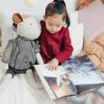 Child reading a picture book with a stuffed toy in a cozy bedroom setting.