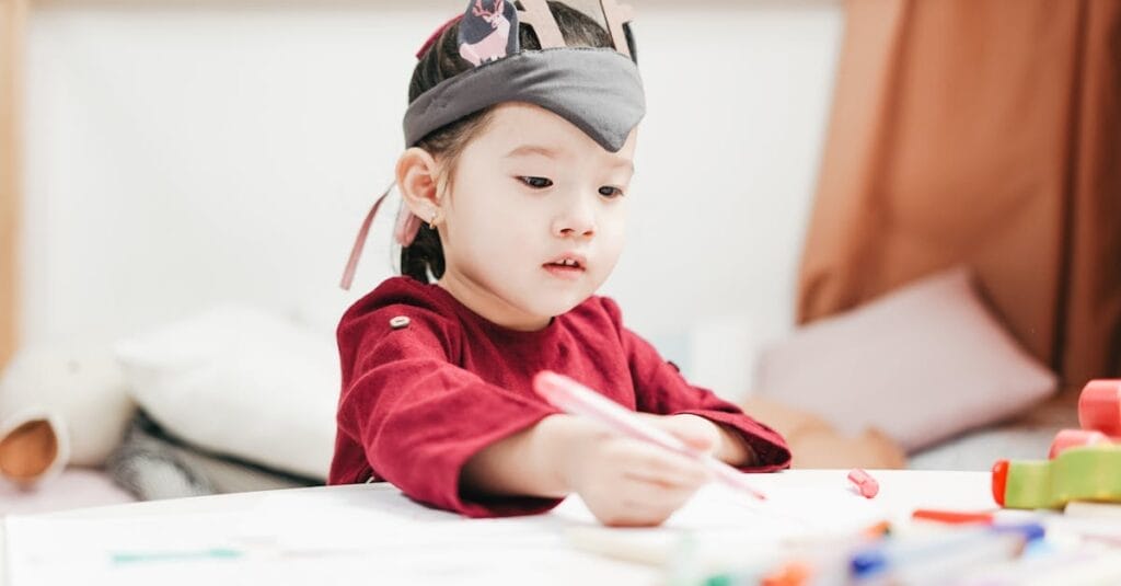 A young child wearing a headpiece enjoying drawing in an indoor playroom setting.