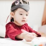 A young child wearing a headpiece enjoying drawing in an indoor playroom setting.